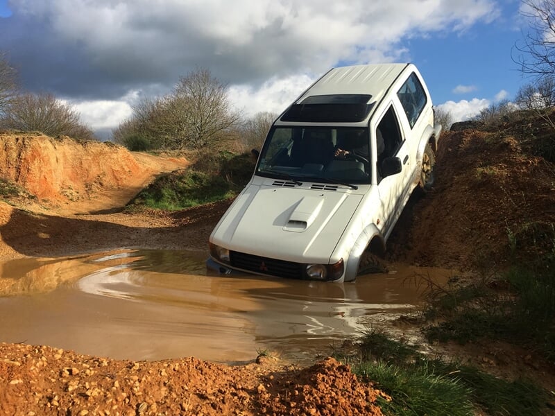 Un 4x4 blanc s'enfonce dans une flaque boueuse sur un terrain accidenté en pleine nature.