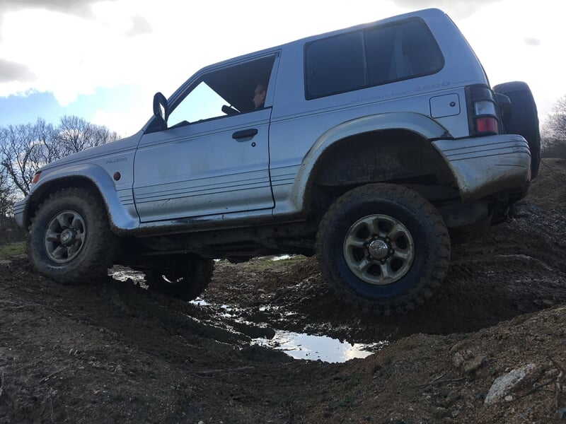 Un 4x4 gris franchit un obstacle boueux sur un terrain tout-terrain avec des arbres en arrière-plan.