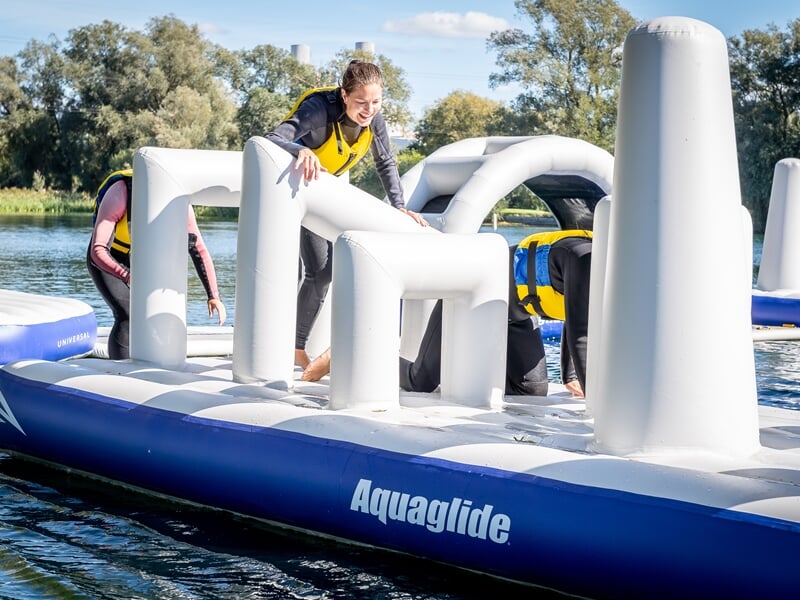 Enfants jouent sur un parcours gonflable aquatique bleu et blanc flottant sur un lac sous un ciel clair.