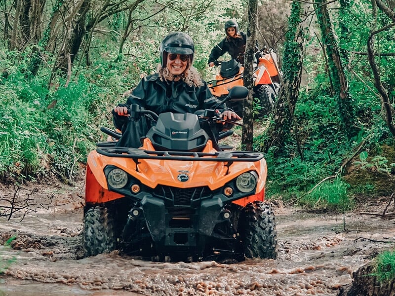 Personne souriante conduit un quad orange sur un chemin boueux en forêt dense.
