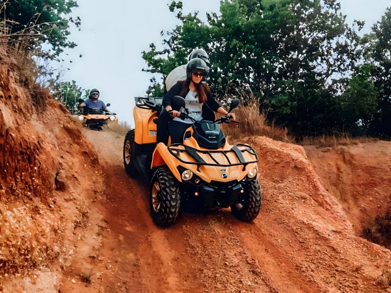 Un homme descend une pente rouge en quad orange dans un paysage naturel avec des arbres verts.