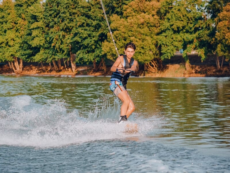 Un homme en gilet de sauvetage fait du wakeboard sur un lac, créant des éclaboussures d'eau.