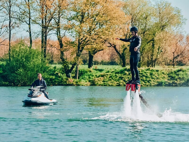Un homme en flyboard sur un lac calme, entouré d'arbres verts sous un ciel bleu clair.