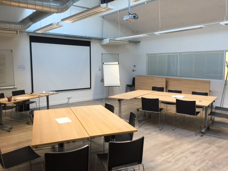 Grande salle de réunion moderne avec tables en bois, chaises noires, écran de projection et tableau blanc.