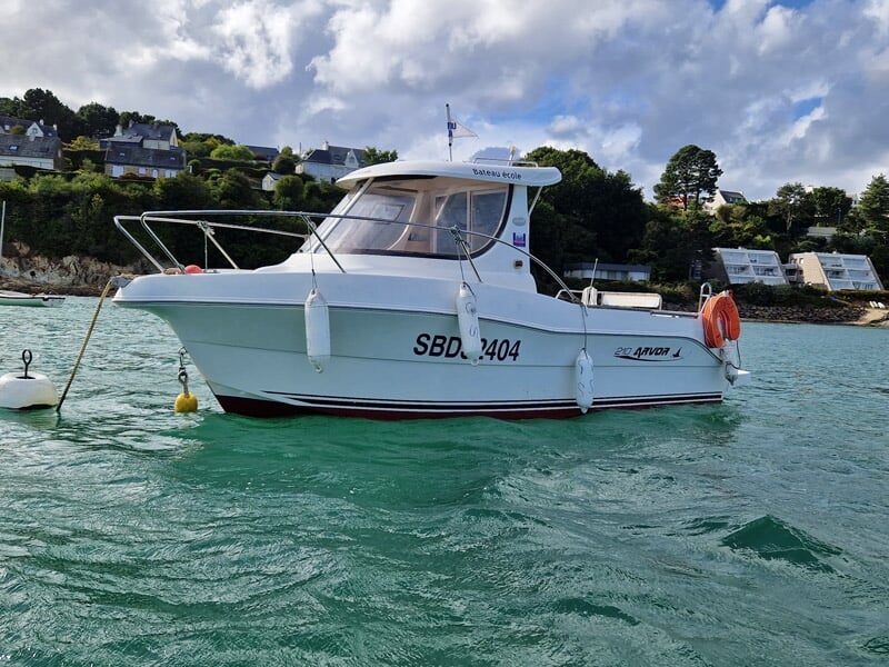 Bateau école Arvor 210 amarré proche du rivage, coque blanche, mer verte, ciel nuageux et colline bordée de maisons bretonnes.