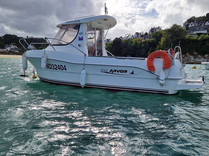 Bateau à moteur blanc Arvor 210 vu depuis l’arrière, bouée de sauvetage orange fixée à la rambarde, eau transparente et côte résidentielle en arrière-plan.