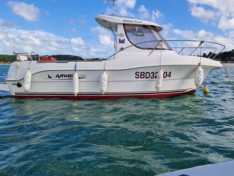 Vue latérale d’un bateau école Arvor 210 amarré, pare-battages suspendus sur la coque, mer calme et paysage côtier boisé en arrière-plan.