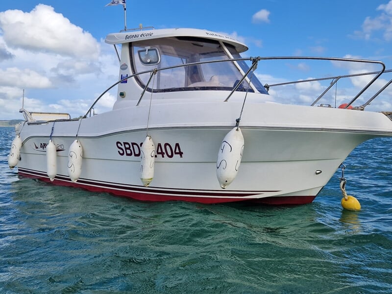 Bateau école blanc de type Arvor 210 flottant sur une mer turquoise, équipé de pare-battages, amarré à une bouée jaune, ciel bleu parsemé de nuages.