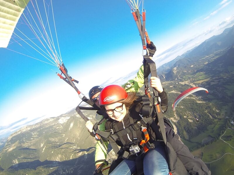 Personne en parapente en tandem avec un instructeur, survolant un paysage montagneux sous un ciel bleu clair.