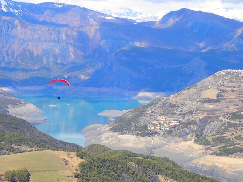 Parapentiste volant au-dessus d'un grand lac entouré de montagnes avec un ciel partiellement nuageux.