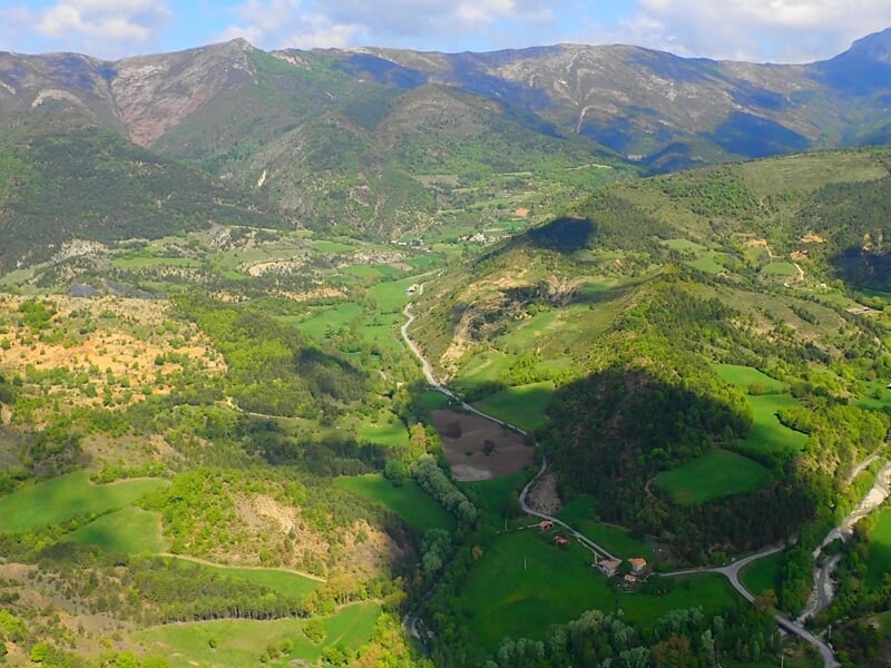 Vue aérienne d'une vallée verdoyante avec des collines, des routes et des champs sous un ciel clair.