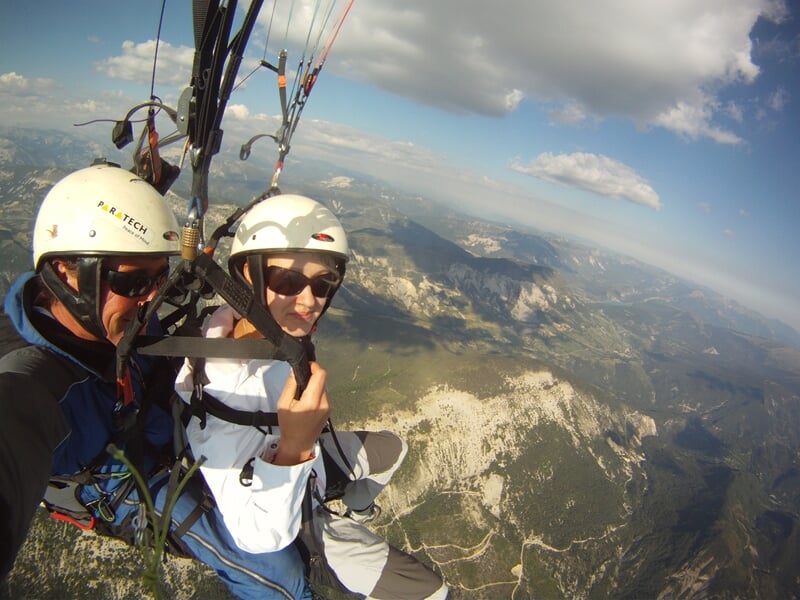 Deux personnes en parapente en tandem survolant un paysage montagneux avec un ciel bleu et quelques nuages.