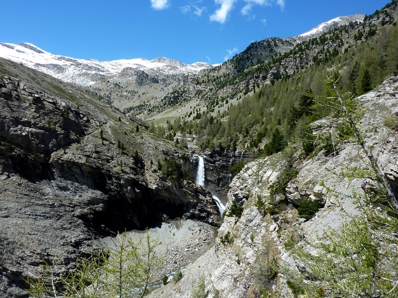Paysage montagneux avec une cascade d'eau, des arbres verts et des sommets enneigés en arrière-plan.