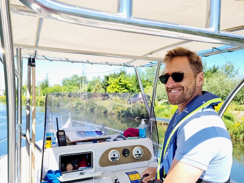 Homme souriant portant un gilet de sauvetage debout à la barre d'un bateau sur une rivière bordée d'arbres.