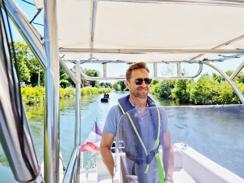 Homme souriant avec lunettes de soleil et gilet de sauvetage debout sur un bateau naviguant sur une rivière calme.