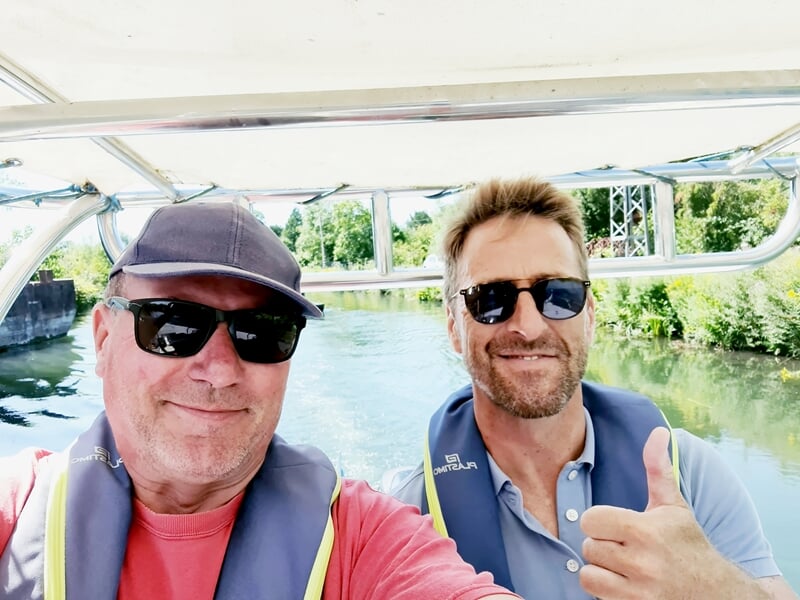 Deux hommes souriants portant des lunettes de soleil sur un bateau, l'un fait un pouce levé, rivière et pont en arrière-plan.