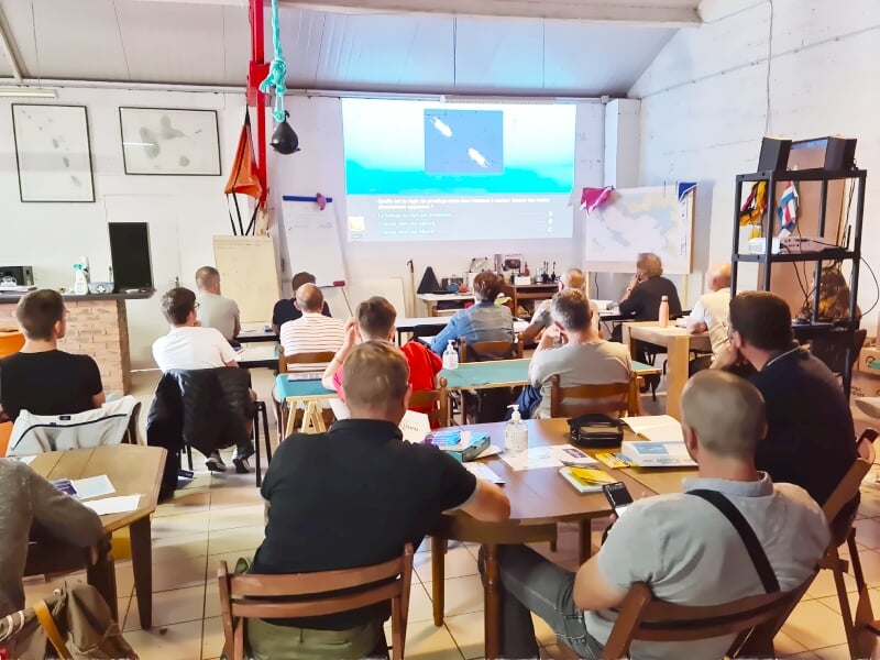 Salle de classe avec plusieurs personnes assises regardant un écran de projection, décorée avec des posters maritimes.