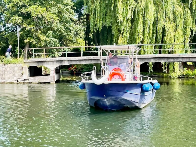 Petit bateau bleu avec des bouées bleues naviguant sur une rivière calme sous un pont, entouré de verdure.