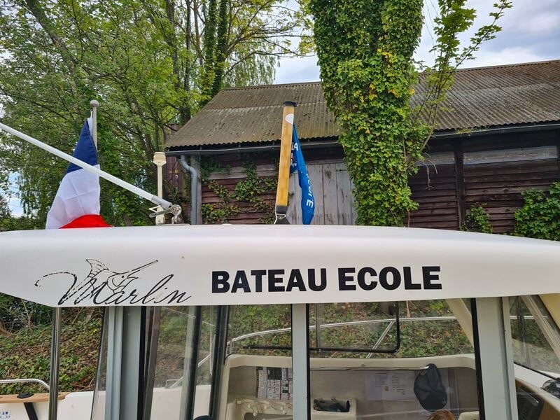 Gros plan sur l'inscription 'BATEAU ECOLE' sur un bateau avec un drapeau français et un environnement boisé.