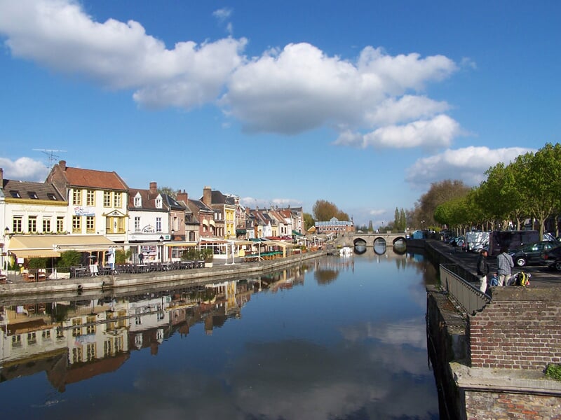 Vue d'un canal calme bordé de maisons anciennes et d'arbres sous un ciel bleu avec quelques nuages blancs.