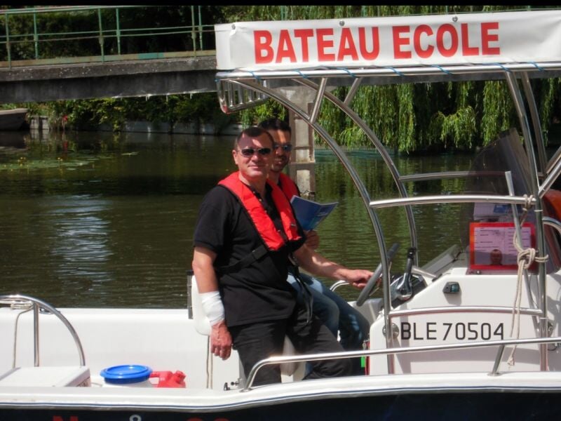 Deux hommes souriants sur un petit bateau-école amarré près d'un pont, entouré d'eau et de verdure.