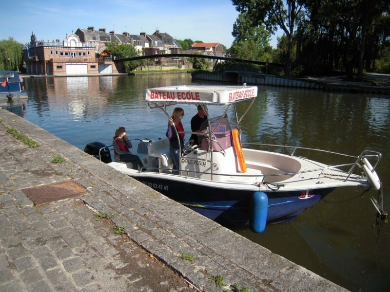 Petit bateau-école amarré le long d'un quai en pierre avec trois personnes à bord sous un ciel ensoleillé.