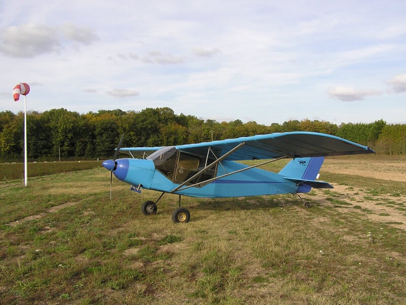 Avion ultraléger bleu stationné sur un terrain herbeux avec un mât de vent et une forêt en arrière-plan.
