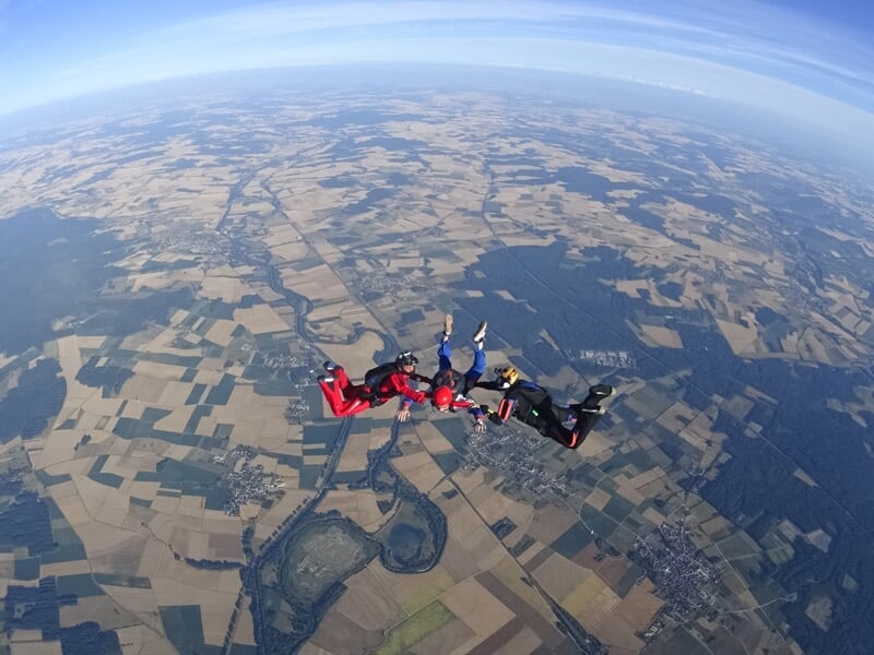 Quatre parachutistes en formation en cercle au-dessus d'un paysage rural avec champs et forêts visibles.