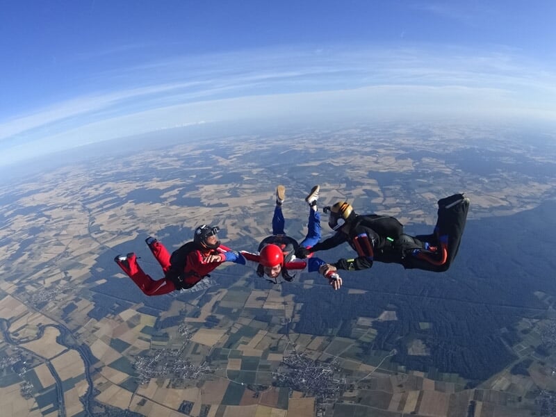 Quatre parachutistes en formation en cercle, volant en chute libre au-dessus d'un paysage agricole étendu.