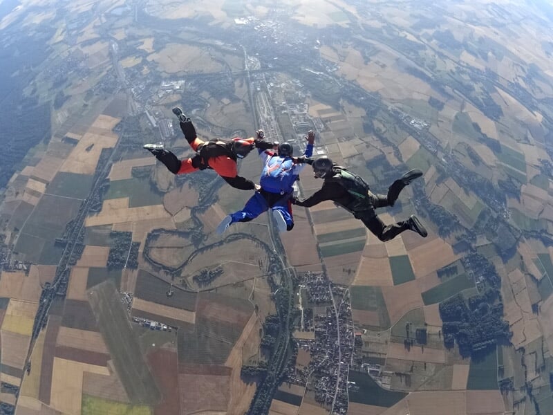 Quatre parachutistes en formation en cercle, volant en chute libre au-dessus d'un paysage de campagne avec rivières.