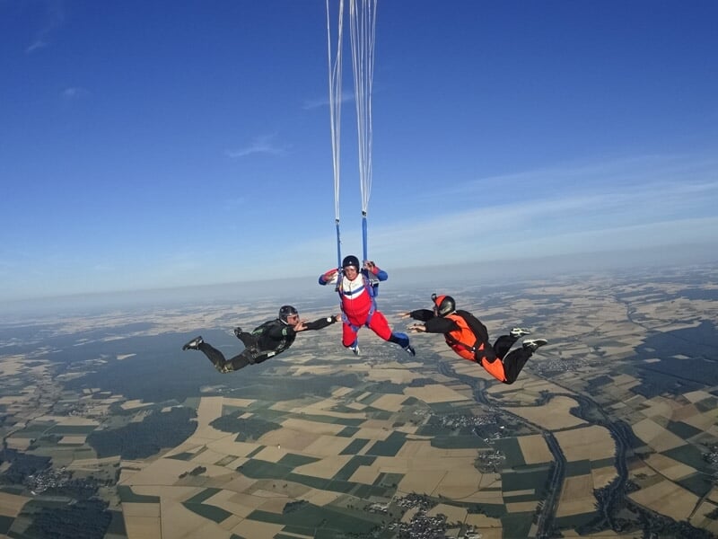 Trois parachutistes en chute libre entourant un parachutiste en combinaison rouge au-dessus d'un paysage rural.