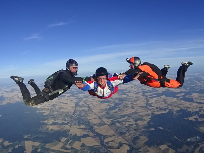 Trois parachutistes en formation en chute libre, tenant les mains au-dessus d'un paysage de champs et forêts.