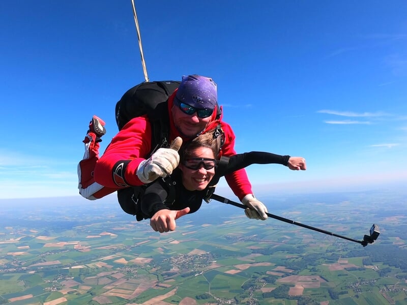 Expérience de parachutisme en tandem avec un instructeur vêtu de rouge et son élève en pleine chute libre. Les deux affichent un large sourire, le pouce levé, tandis qu'une perche à selfie capture leu