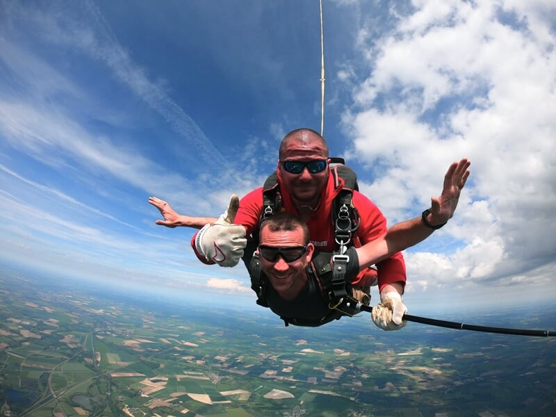 Saut en parachute en tandem sous un ciel bleu avec des nuages. L'instructeur en rouge et son élève, tous deux équipés de lunettes de protection, profitent de la chute libre avec un pouce levé en signe