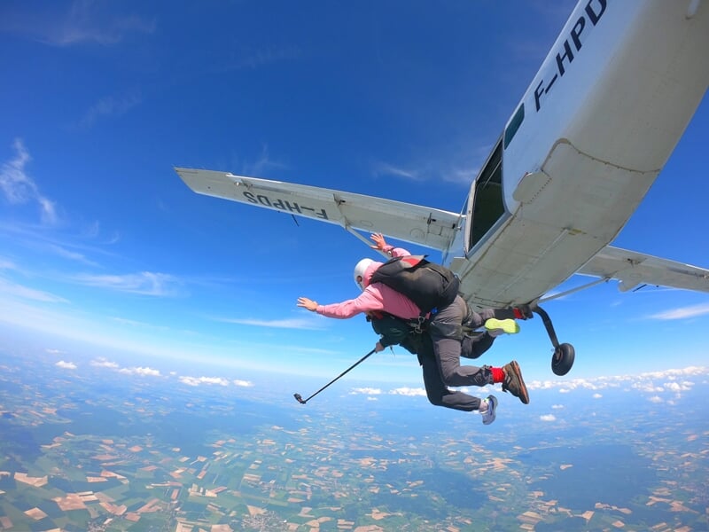 Moment d'adrénaline lors d'un saut en parachute tandem depuis un avion en plein ciel. L'instructeur et son élève, attachés ensemble, s'élancent dans le vide au-dessus d'un paysage verdoyant et agricol