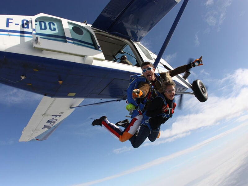 Instant de pure adrénaline lors d'un saut en parachute tandem depuis un avion bleu et blanc. L'instructeur et son élève, visiblement excités, plongent dans le vide avec enthousiasme sous un ciel dégag