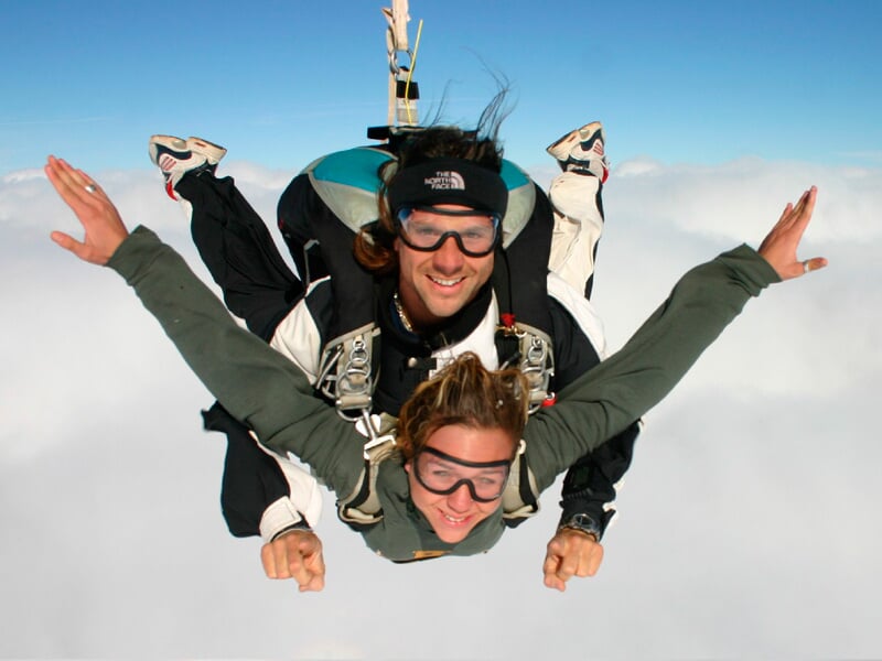 Saut en parachute tandem au-dessus d'une mer de nuages. L'instructeur et son élève, souriants et équipés de lunettes de protection, profitent de la chute libre avec les bras écartés, en pleine sensati