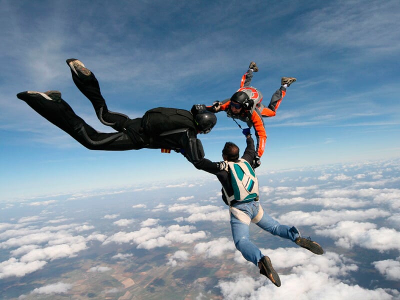 Trois parachutistes en chute libre se tiennent par les mains au-dessus d'un paysage nuageux et terrestre.