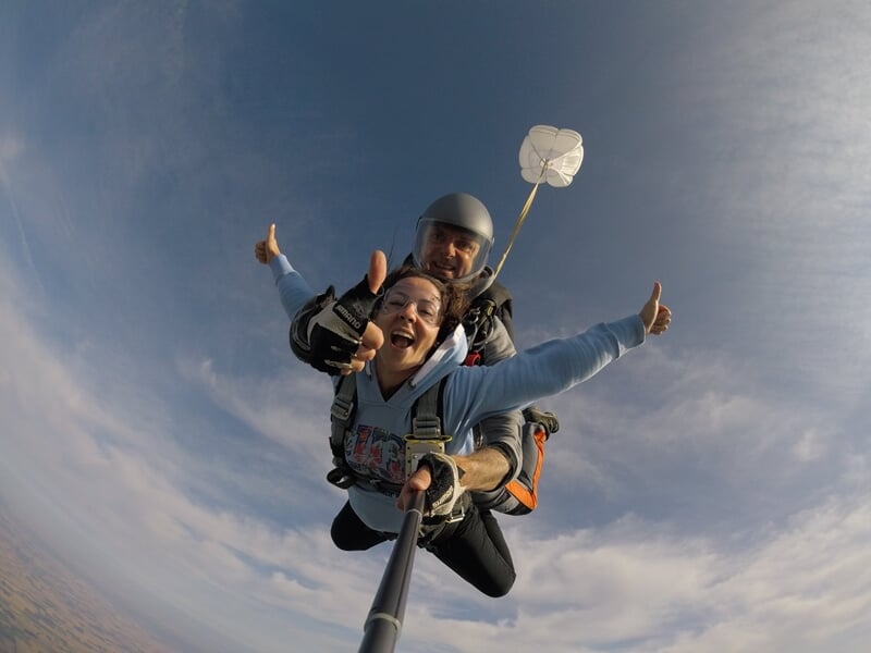 Saut en parachute en tandem, selfie avec perche, ciel bleu et petit parachute de secours blanc.