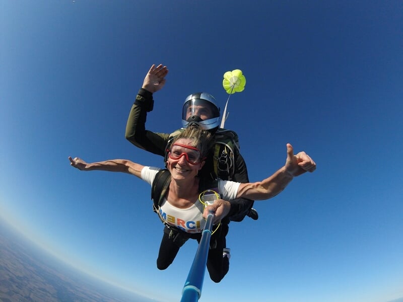 Parachutiste en tandem souriant, selfie avec perche, ciel bleu clair et petit parachute de secours jaune.