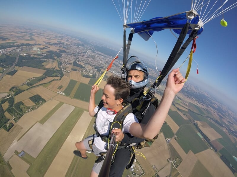 Deux parachutistes en tandem, selfie avec perche, survolant des champs et villages sous un ciel clair.