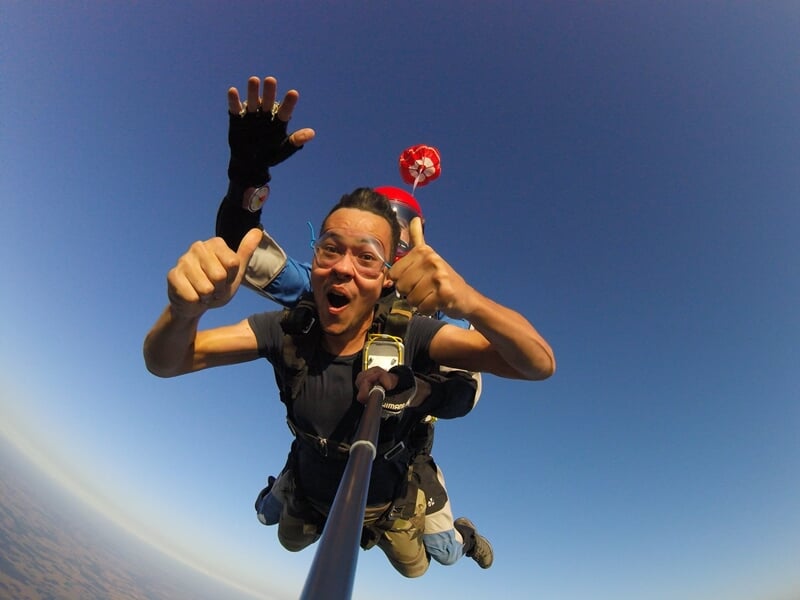 Homme en chute libre en tandem, selfie avec perche, ciel bleu et petit parachute de secours rouge visible.