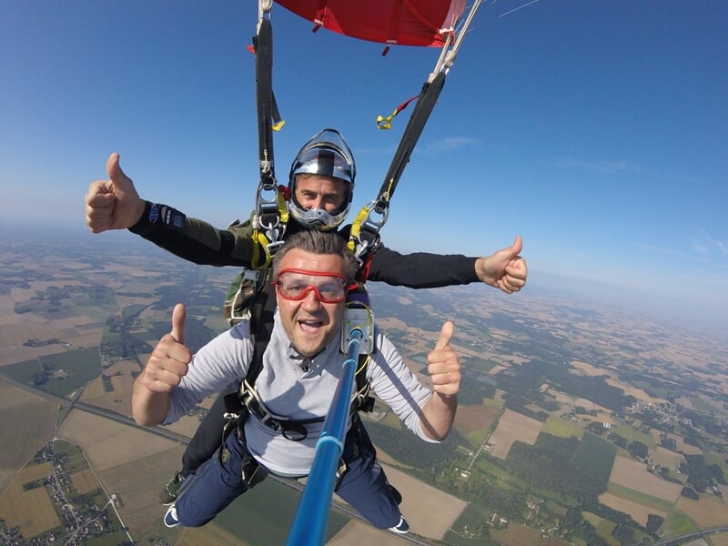 Deux parachutistes en tandem souriants, selfie avec perche, parachute rouge ouvert et campagne en arrière-plan.