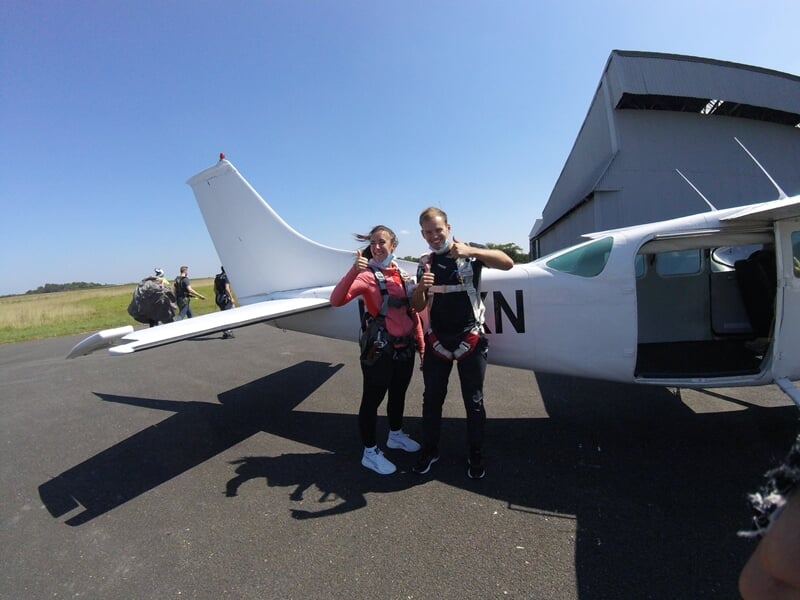 Deux personnes en tenue de saut posant devant un petit avion blanc sur une piste d'aérodrome ensoleillée.