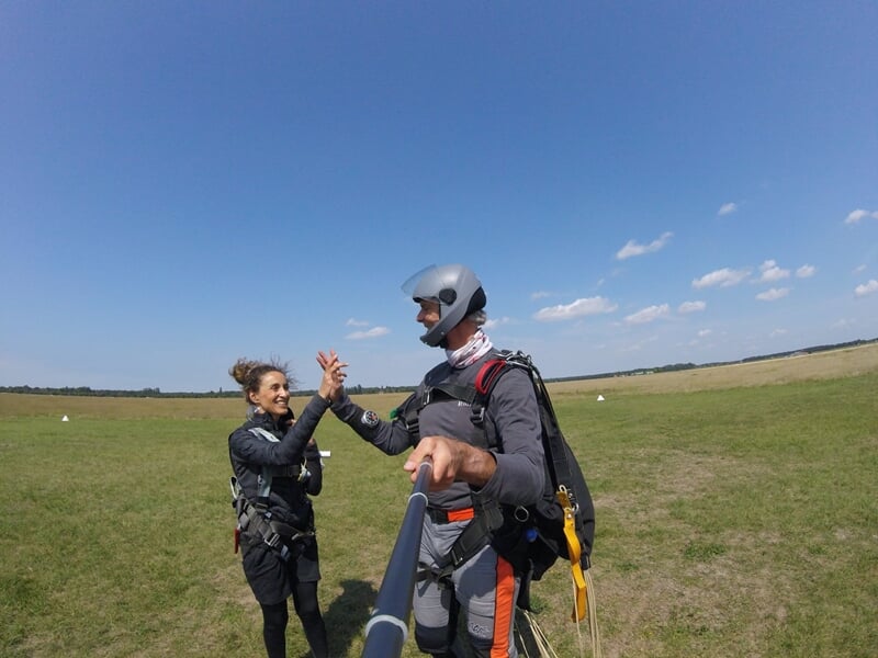 Deux parachutistes en tenue de saut se saluant sur une pelouse verte à l'aérodrome, ciel bleu clair.