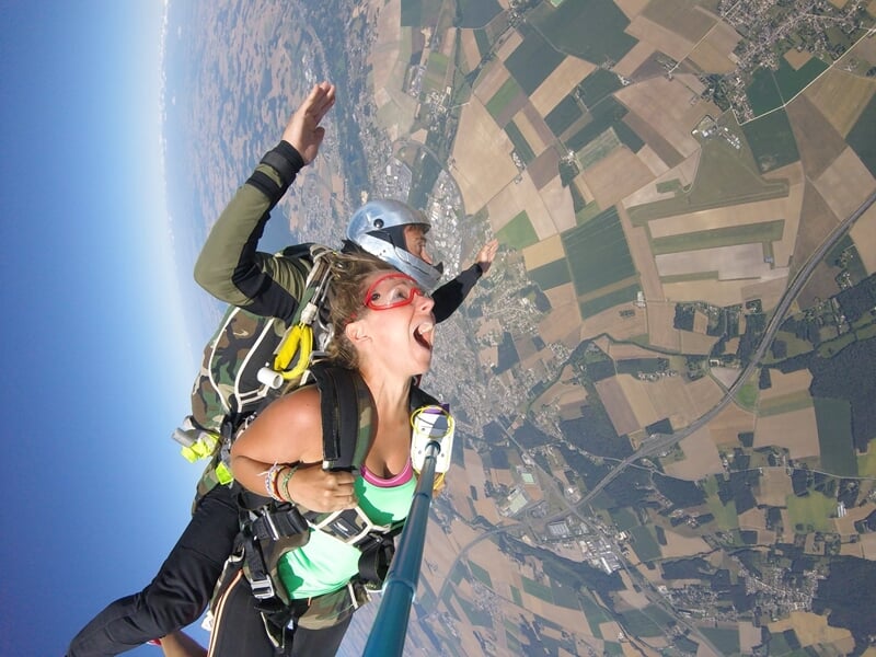 Parachutiste en tandem en chute libre, selfie avec perche, survolant des champs et villages sous un ciel clair.