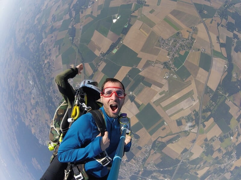 Parachutiste en tandem avec lunettes rouges, selfie avec perche, expression de surprise et campagne en dessous.