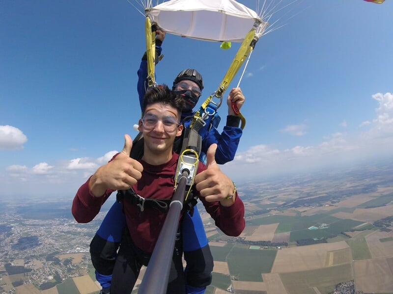 Homme en chute libre en tandem, selfie avec perche, expression de joie intense, paysage rural en arrière-plan.