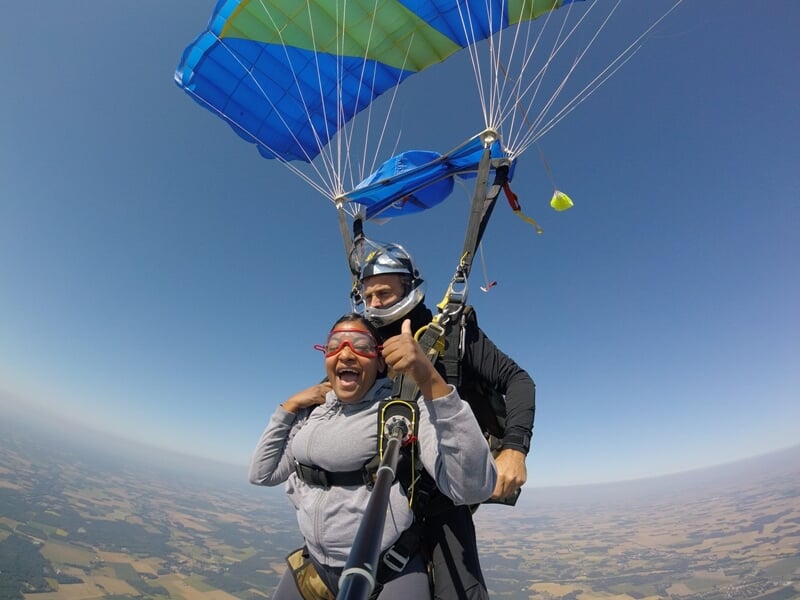 Jeune homme en chute libre en tandem, selfie avec perche, souriant, survolant des champs et villages.