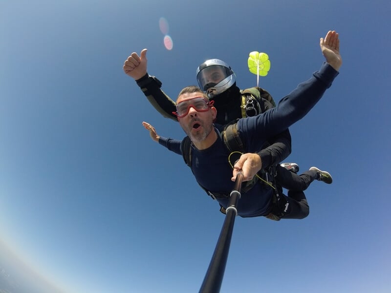 Parachutiste en tandem souriant, selfie avec perche, parachute bleu ouvert, ciel bleu et campagne en dessous.